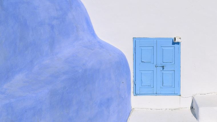 Painted timber shutters and windows on a whitewashed wall in Oia, Santorini, the joinery and glazing that salt air degrades within a few seasons